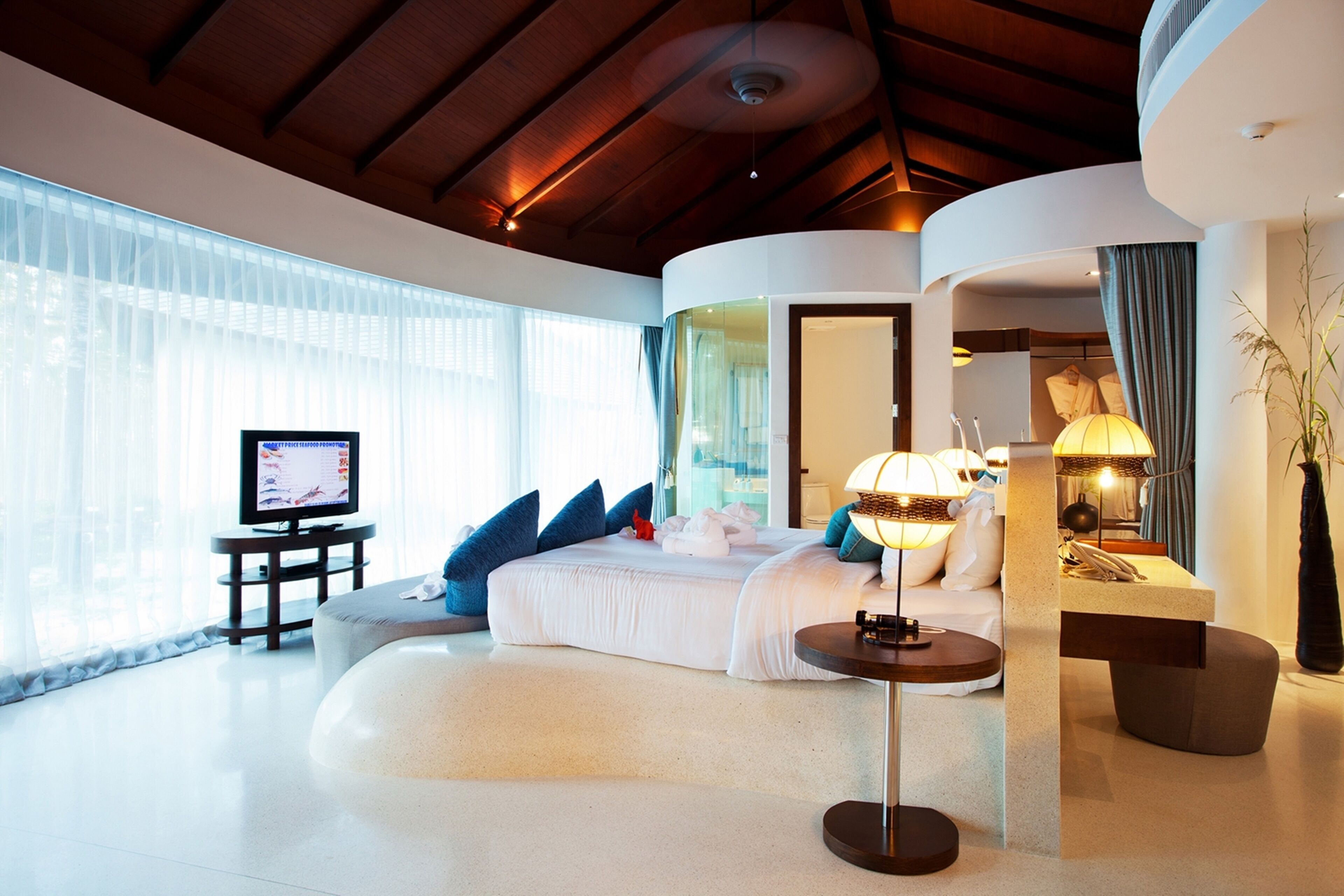 Una habitación de hotel moderna con techo curvo, una cama grande con ropa de cama blanca, un televisor sobre un mueble bajo y una mesita de noche con lámpara.
