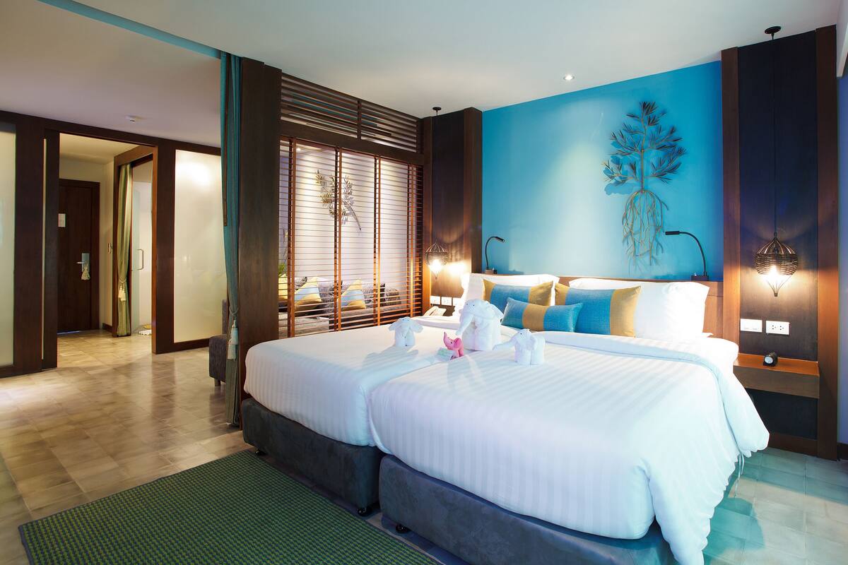 Une chambre d’hôtel avec deux lits, une grande fenêtre avec stores, une œuvre d’art accrochée au mur et une table de chevet avec une lampe.