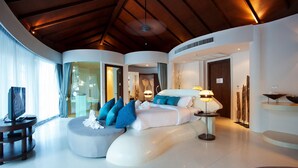 Une chambre d’hôtel moderne avec un plafond incurvé, un grand lit avec une literie bleue et blanche, un coin salon circulaire et un téléviseur posé sur un meuble.