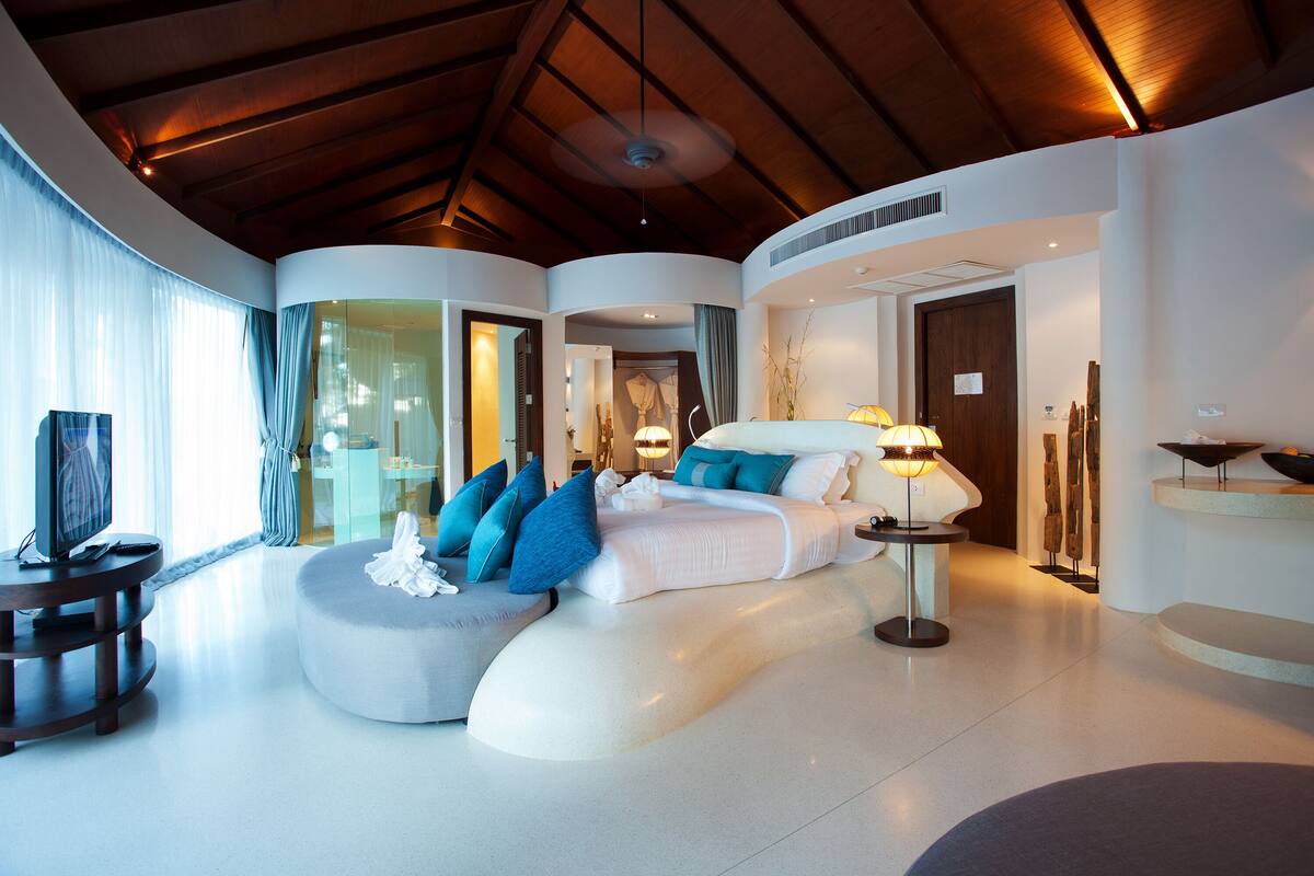 Une chambre d’hôtel moderne avec un plafond incurvé, un grand lit avec une literie bleue et blanche, un coin salon circulaire et un téléviseur posé sur un meuble.