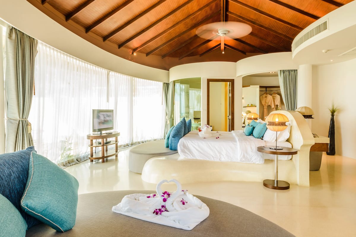 Une chambre d’hôtel moderne dotée d’un plafond incurvé, d’un grand lit avec du linge de maison blanc, d’un coin salon avec un canapé et d’un téléviseur posé sur une table d’appoint.
