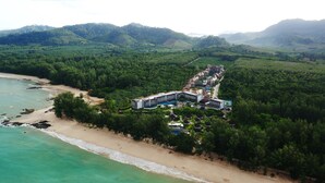 Une vue aérienne d’un complexe hôtelier avec une plage, des collines densément boisées et un long bâtiment doté de nombreux balcons.