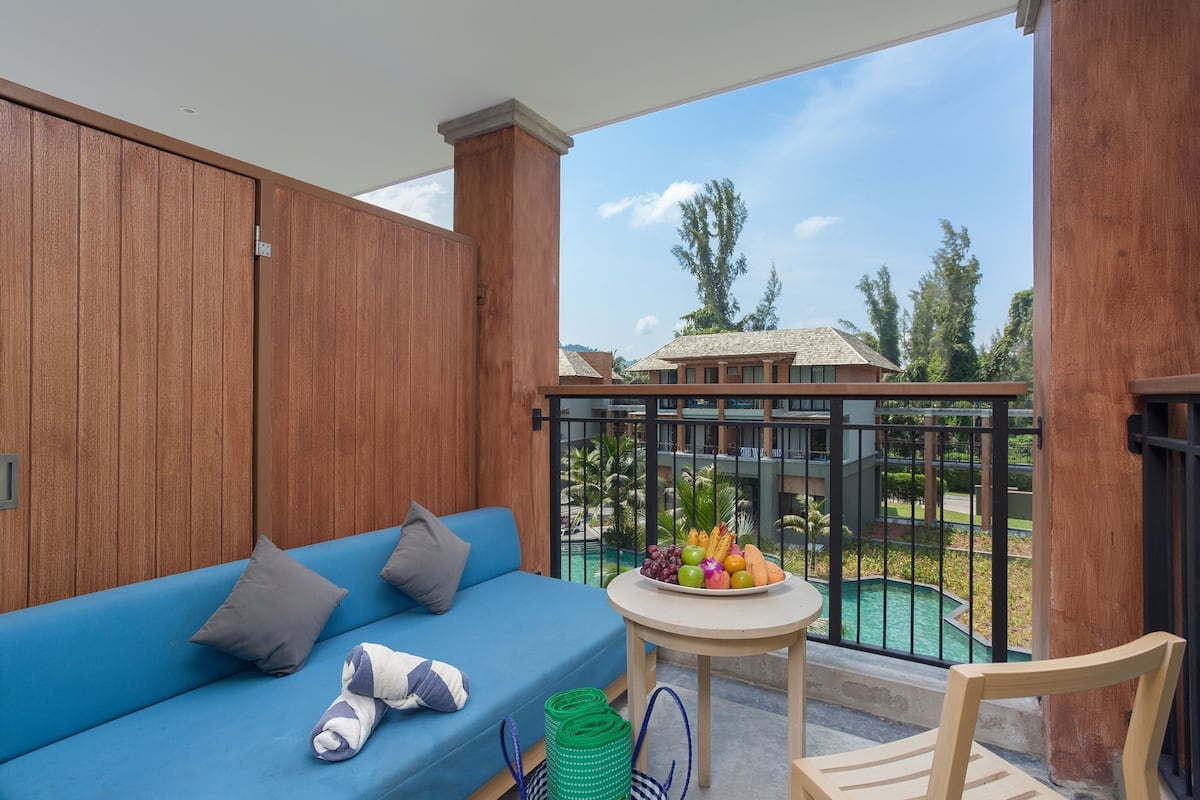 Un balcon avec un canapé bleu, une petite table sur laquelle sont disposés des fruits, et une vue sur une piscine et des immeubles.