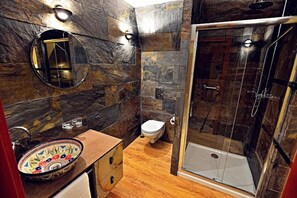 A bathroom with a stone wall, a round mirror, a wooden vanity, a toilet, and a glass-enclosed shower.
