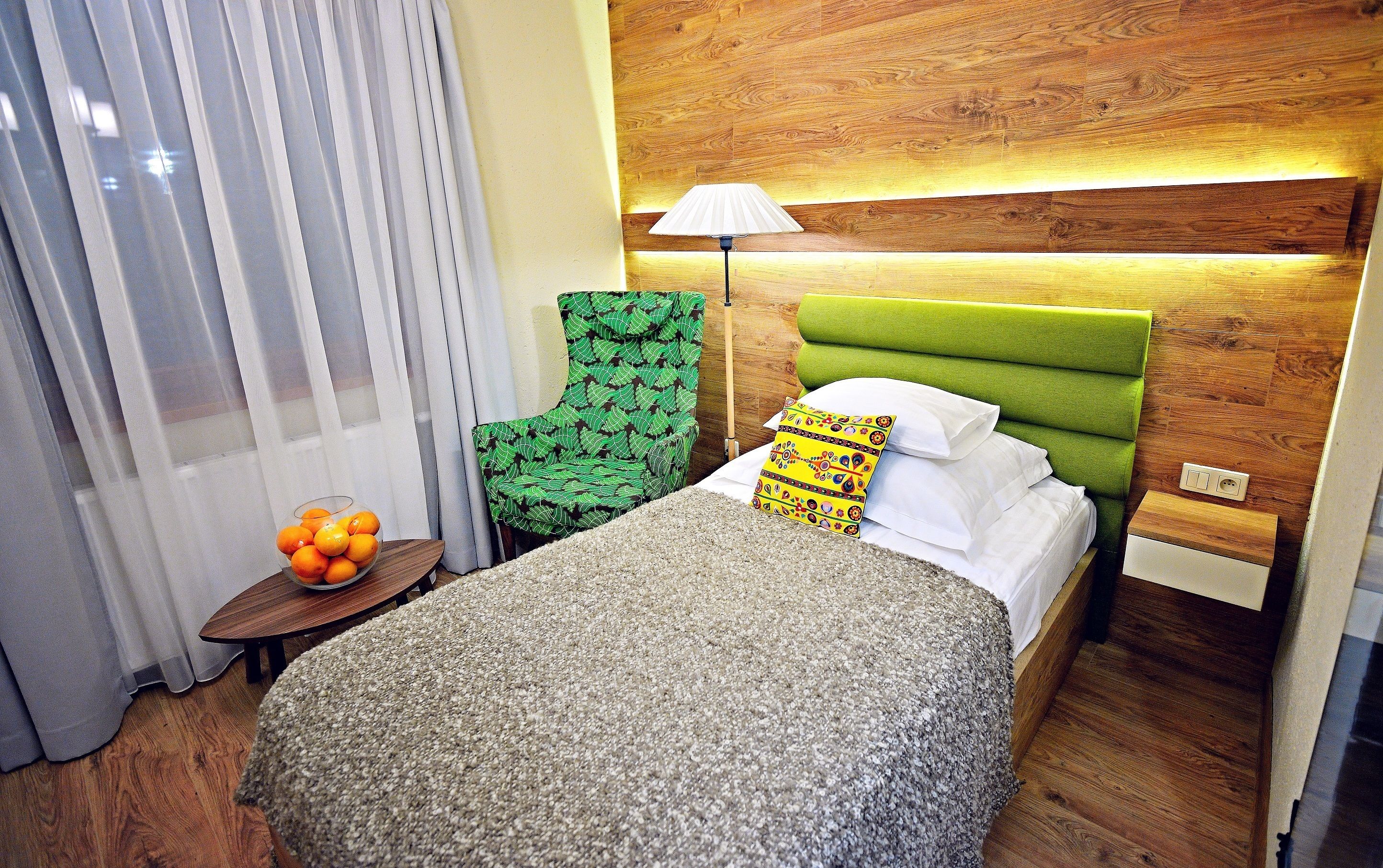 A hotel room with a bed, a green upholstered chair, a lamp, a small table with a bowl of oranges, and a wooden headboard.