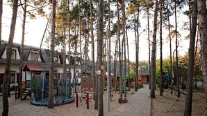 A playground area with a trampoline, climbing structures, and a small slide, surrounded by trees and a brick pathway.