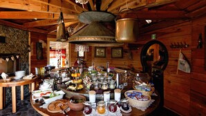 A rustic wooden interior with a central round table laden with various food items and utensils, surrounded by wooden walls and a stone floor.