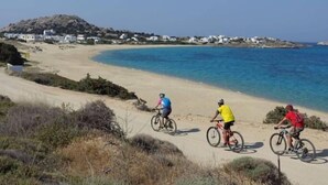 A coastal road with cyclists, clear blue sea, and white buildings in the background. - Mitos Suites (Naxos)