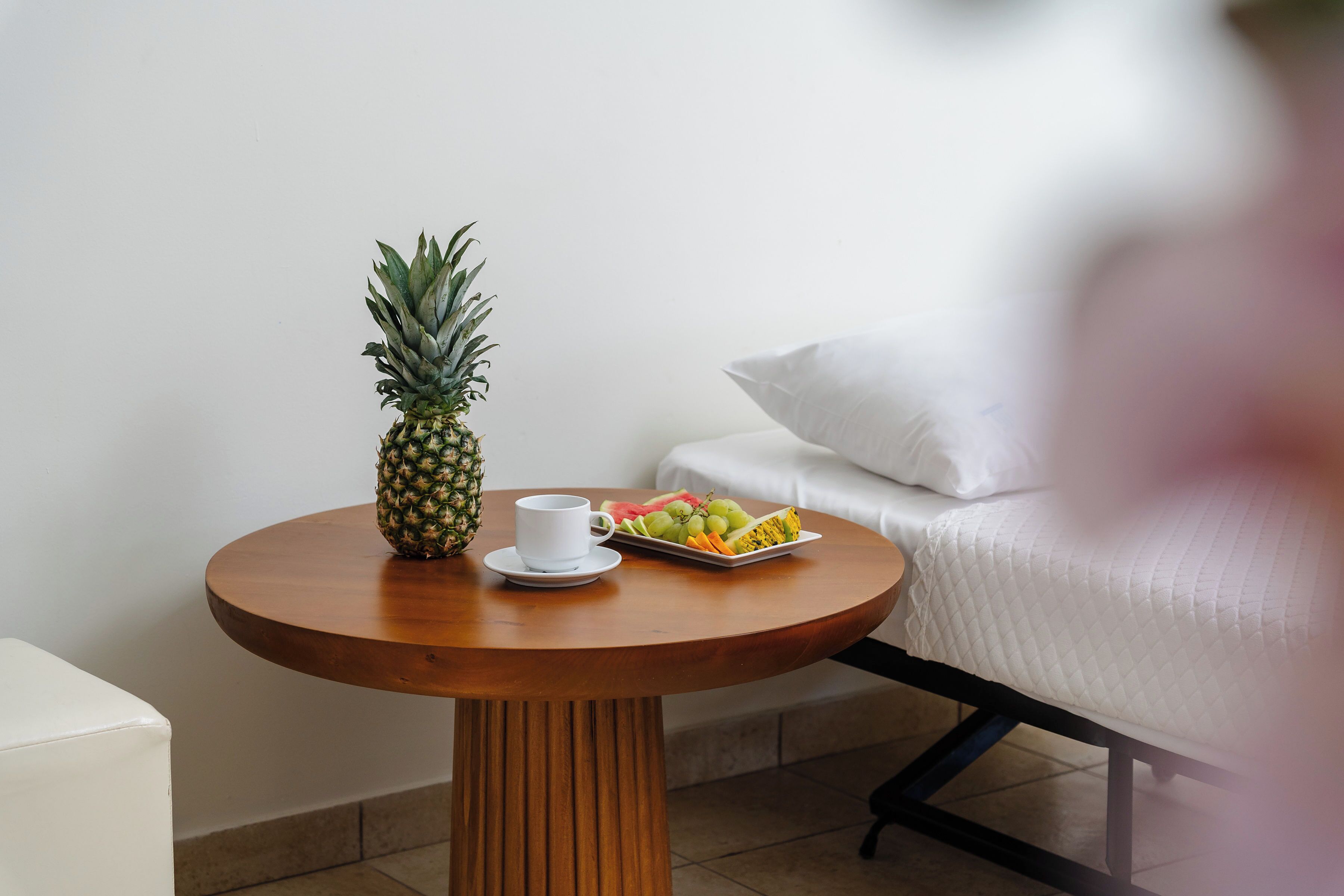 A round wooden table with a pineapple, a white cup, and a tray of fruit in a hotel room.