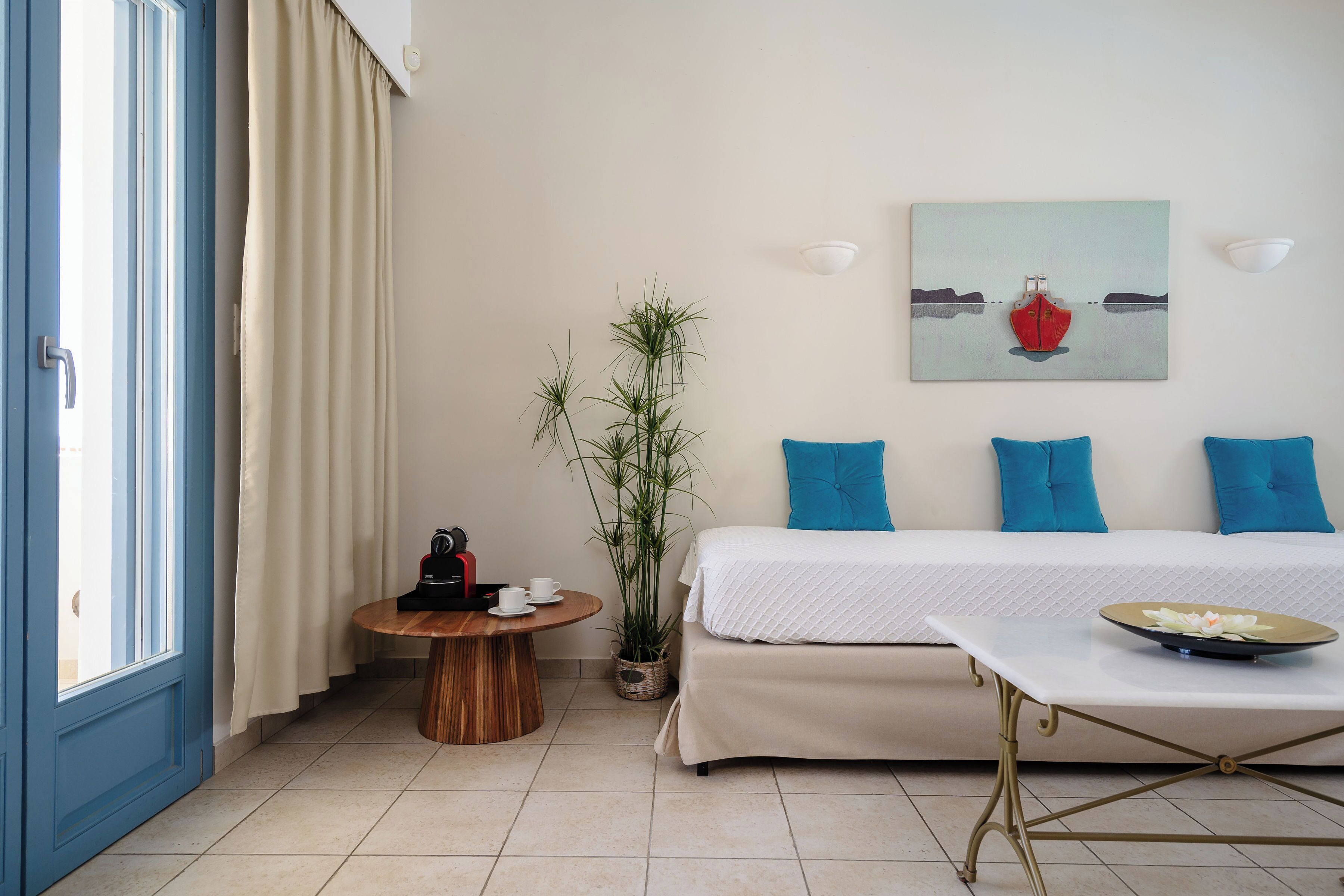 A hotel room with a bed, a round wooden table, a potted plant, and a painting on the wall.