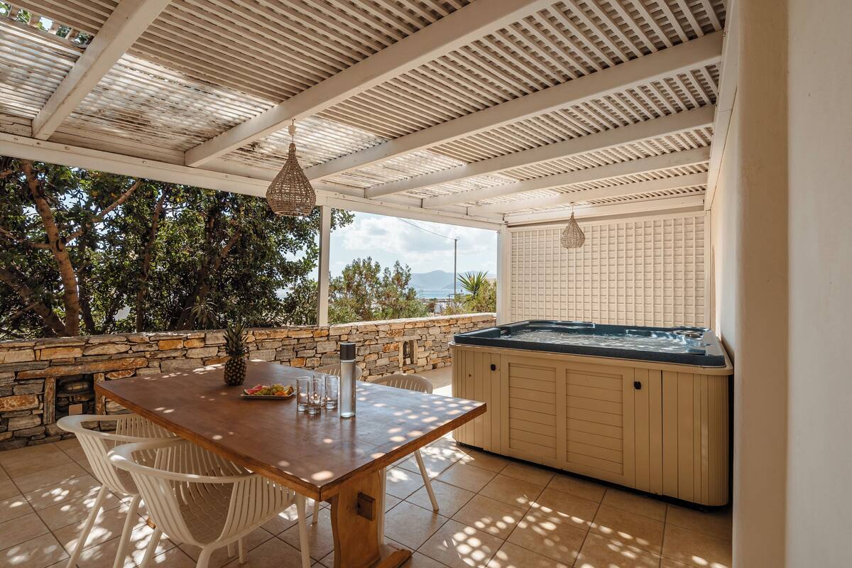 A covered outdoor area with a wooden table, white chairs, a stone wall, and a hot tub.