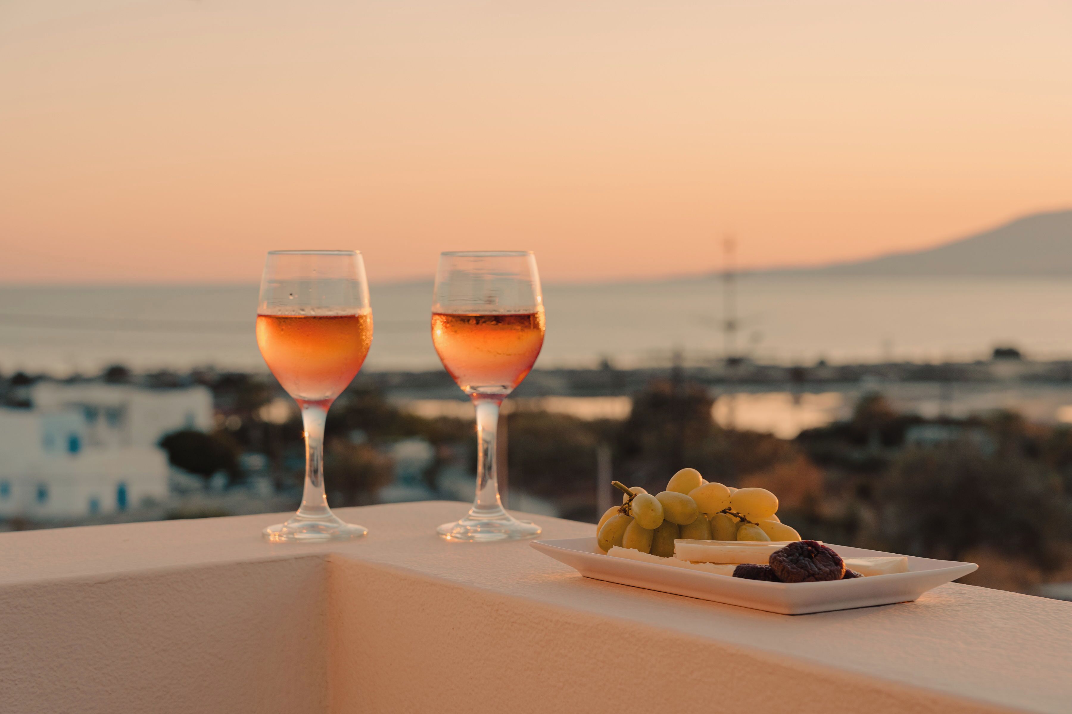 Two glasses of a pink beverage on a balcony with a plate of grapes and a dessert, overlooking a coastal view.