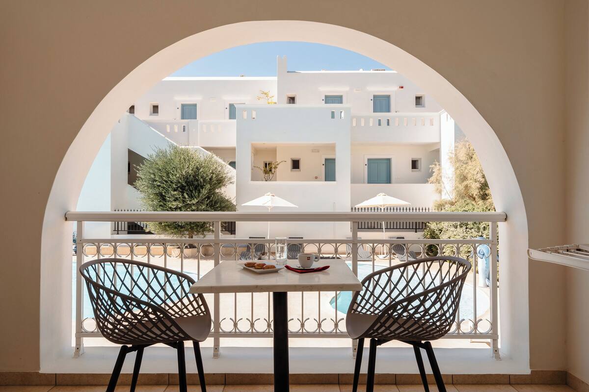 A balcony with a table and two chairs, overlooking a pool and buildings.