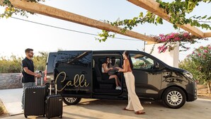 A black van with the text "Calli exceptional transfer" on the side parked under a wooden pergola with pink flowers. - Mitos Suites (Naxos)