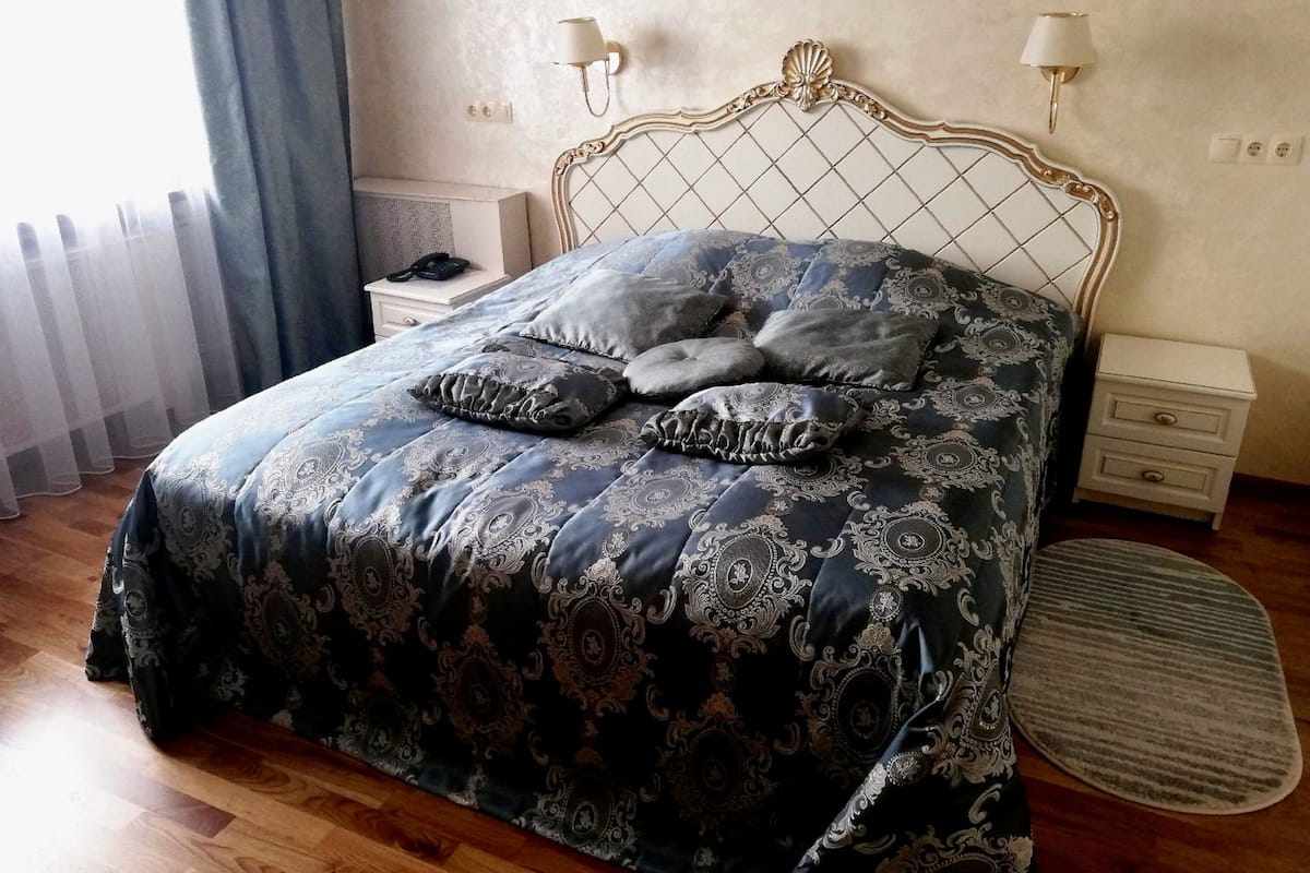 Cama bem arrumada com colcha estampada e almofadas, duas mesas de cabeceira com abajures, um tapete redondo e uma janela com cortinas.