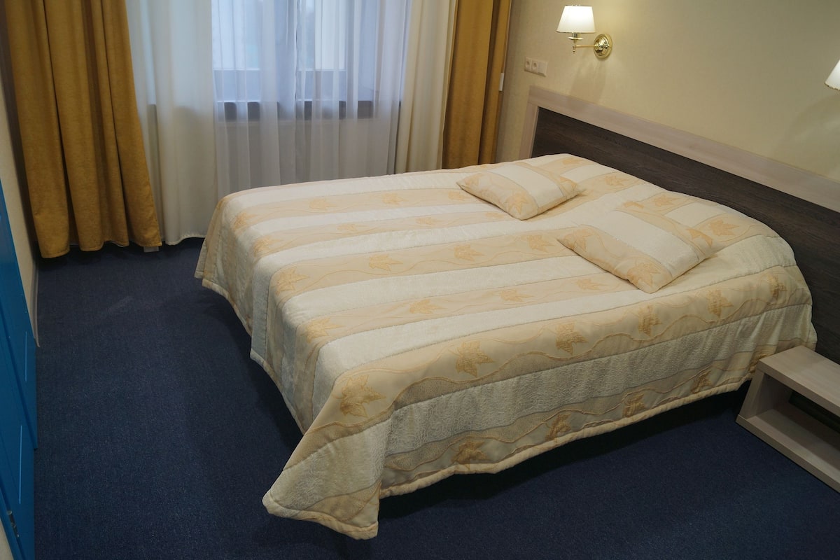 Quarto de hotel com uma cama grande, mesa de cabeceira, luminária e janela com cortinas.