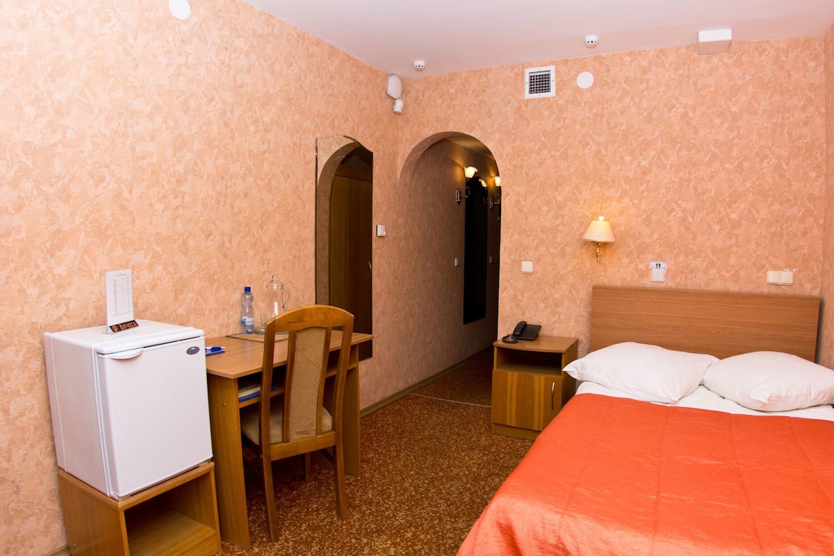 Quarto de hotel com cama, escrivaninha com frigorífico, cadeira, mesa de cabeceira e abajur.