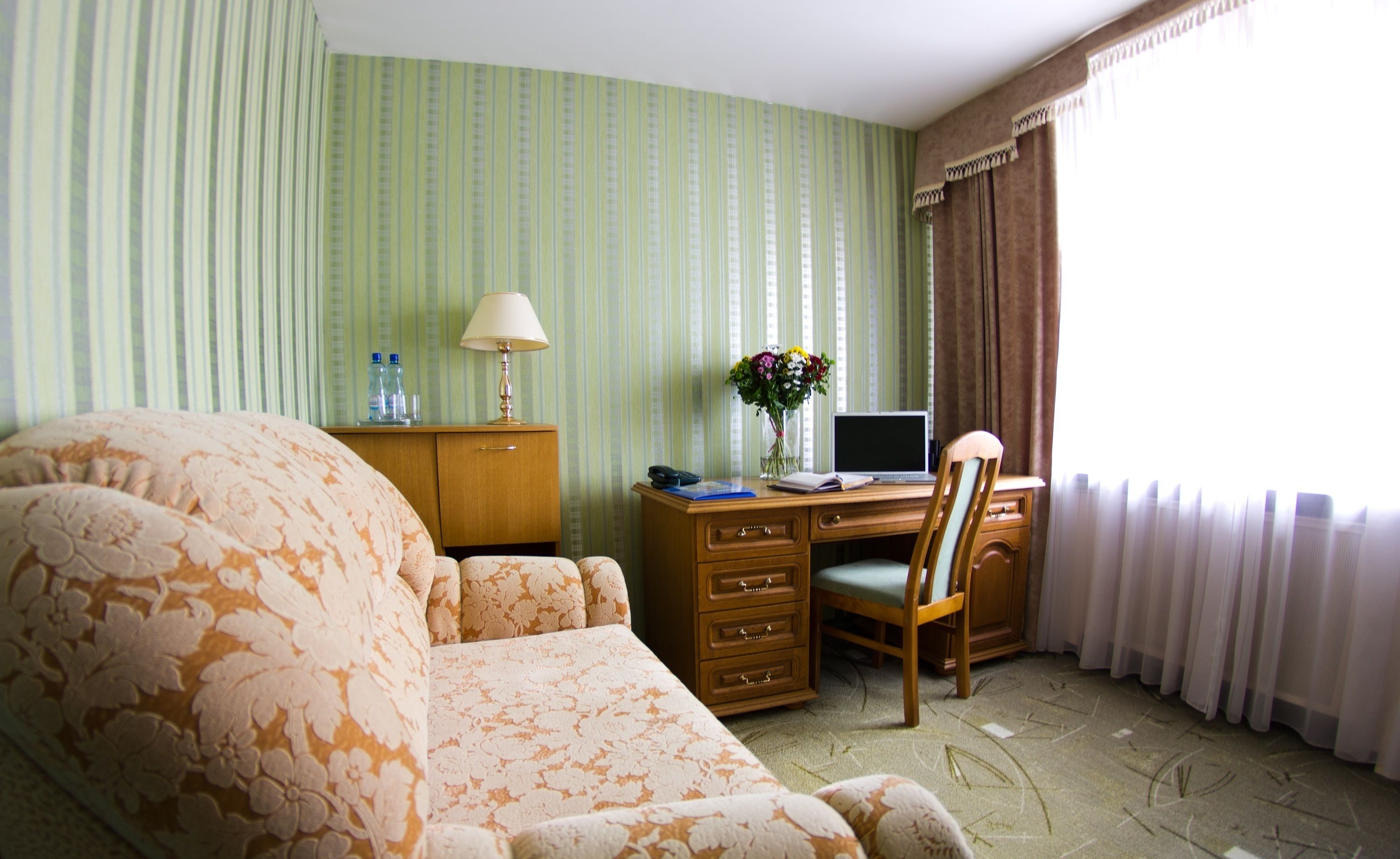 Ein Hotelzimmer mit einem sofa in floralem Muster, einem Holztisch mit einem Laptop, einer Vase mit Blumen und einem Fenster mit Vorhängen.
