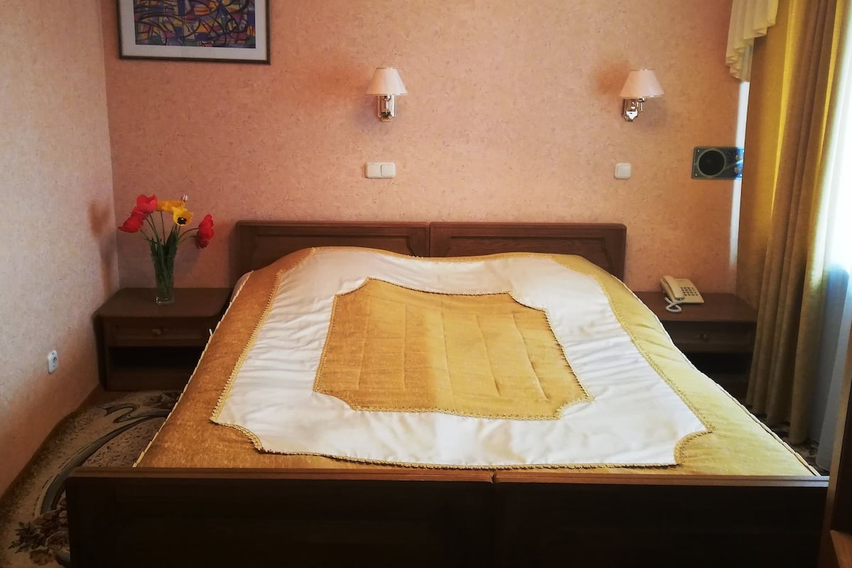 Quarto de hotel com cama, duas luminárias de cabeceira, telefone, quadro decorativo e um vaso com flores.