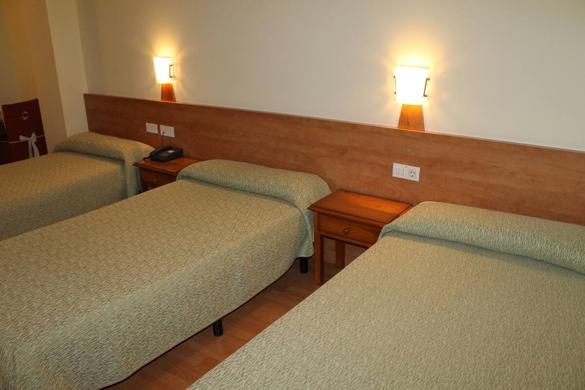 A hotel room with three single beds, each with a wooden headboard and bedside tables. There are two wall-mounted lamps providing light.