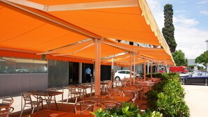 An outdoor cafe area with orange canopies, metal tables, and chairs. - Hotel Flora Parc (Castelldefels)