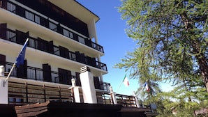 A multi-story hotel with balconies, flags, and a tree in the background. - Baita della Luna (Cesana Torinese)