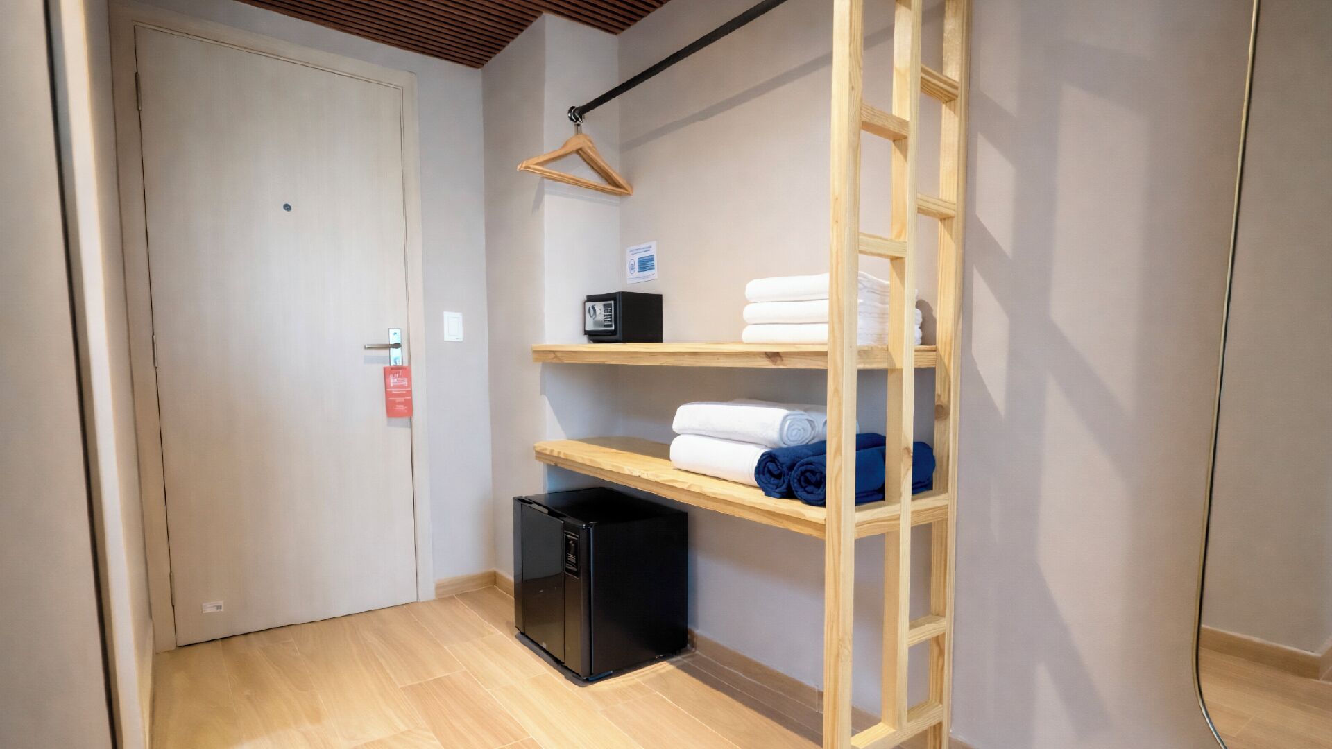 Una camera d'albergo con pavimento in legno, un armadio con ripiani, un cestino nero e una scala in legno.