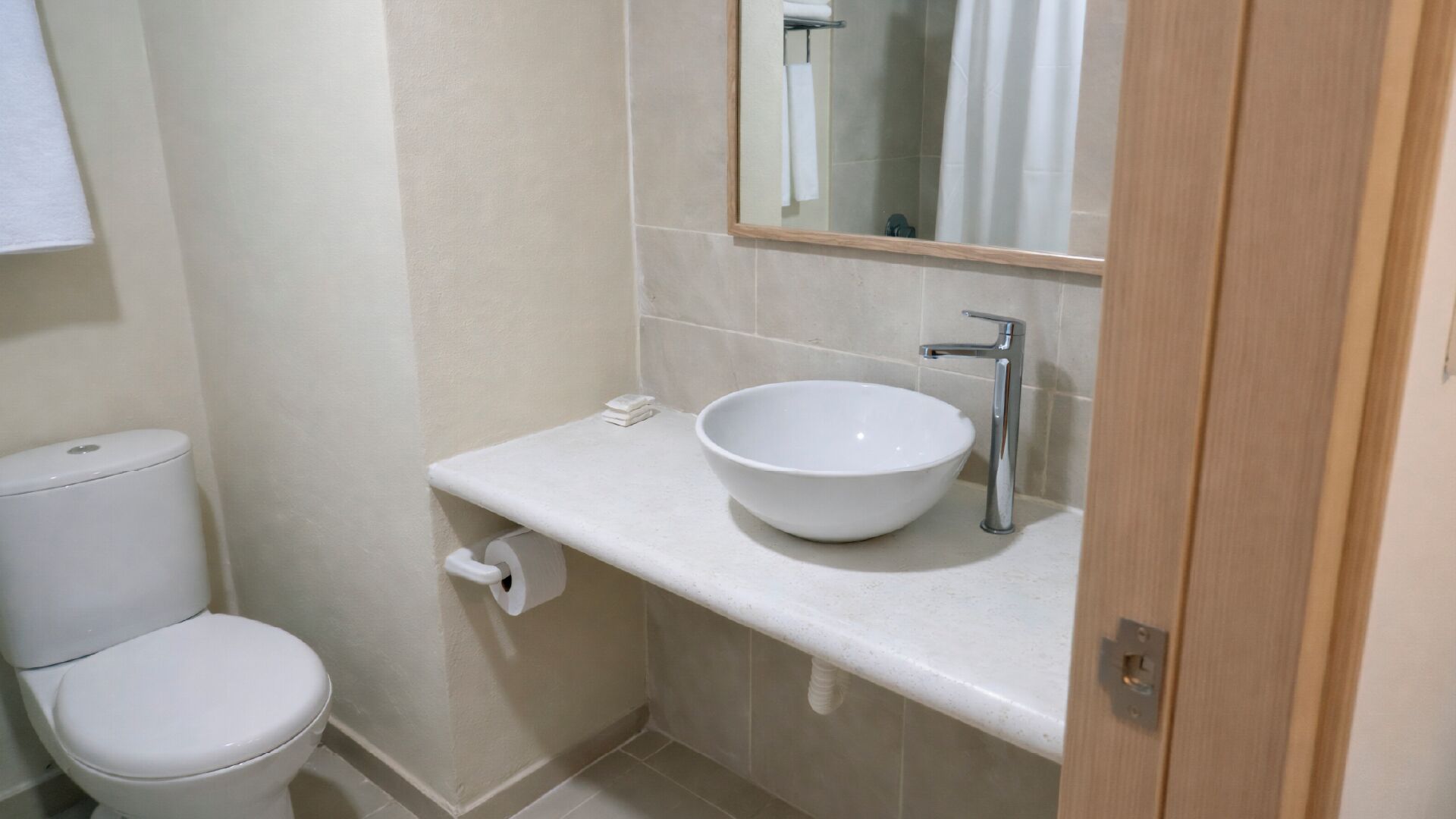 Une petite salle de bain propre, avec des toilettes blanches, un lavabo blanc avec un receveur et un miroir au-dessus du lavabo.