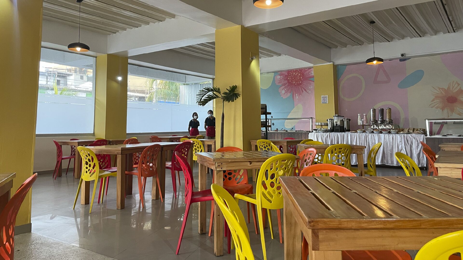 Un comedor con mesas de madera y sillas de plástico de colores, ventanas grandes y un mural en la pared.