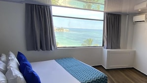 A hotel room with a bed, a window with a view of the sea, and an air conditioning unit.
