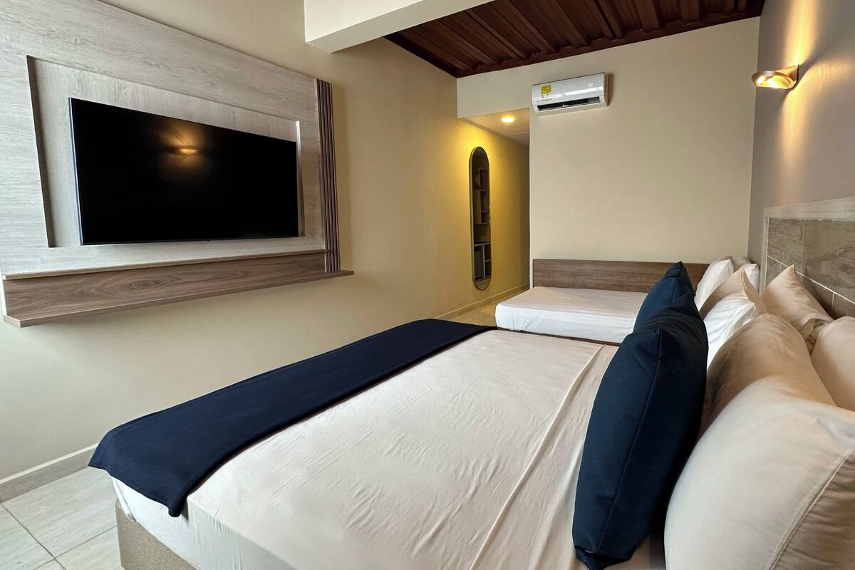 A modern hotel room with a large TV mounted on the wall, a bed with white linens and blue pillows, and a built-in air conditioning unit.