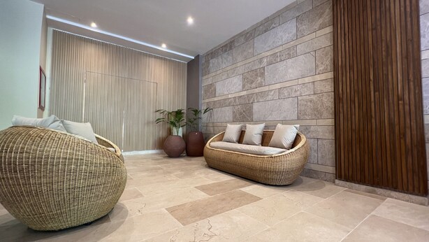 Un área de descanso moderna con un amplio sofá de mimbre, un sillón de mimbre más pequeño y un banco acolchado. Se observan dos plantas grandes en macetas y una pared con textura similar a la piedra.