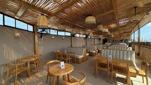 Un restaurante con mobiliario de madera, techo tejido y ventanas amplias.
