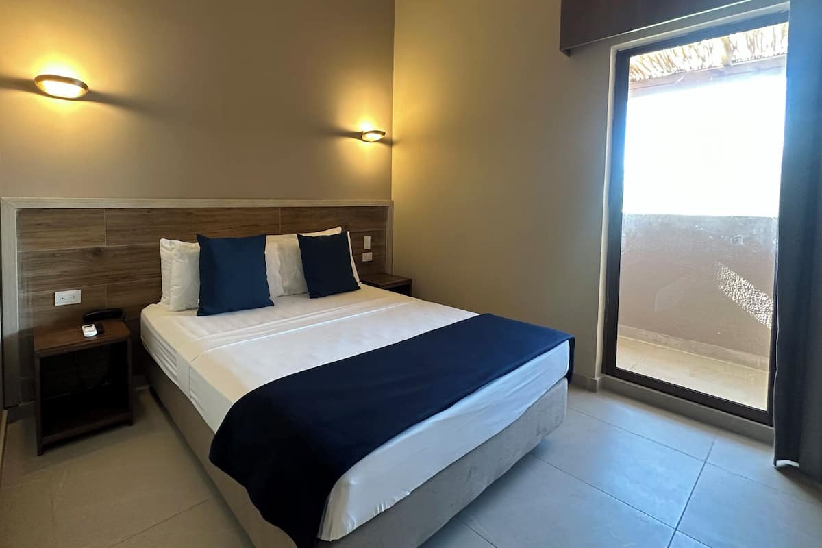 A modern hotel room with a large bed, two bedside tables, a nightstand, and a view of the outdoors through a large window.