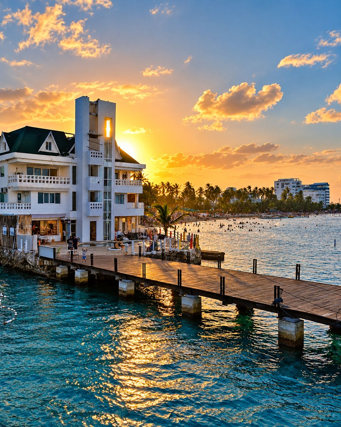 A waterfront hotel with a dock extending into the water, a beach in the background, and a sunset.