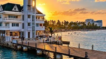 A waterfront hotel with a dock extending into the water, a beach in the background, and a sunset.
