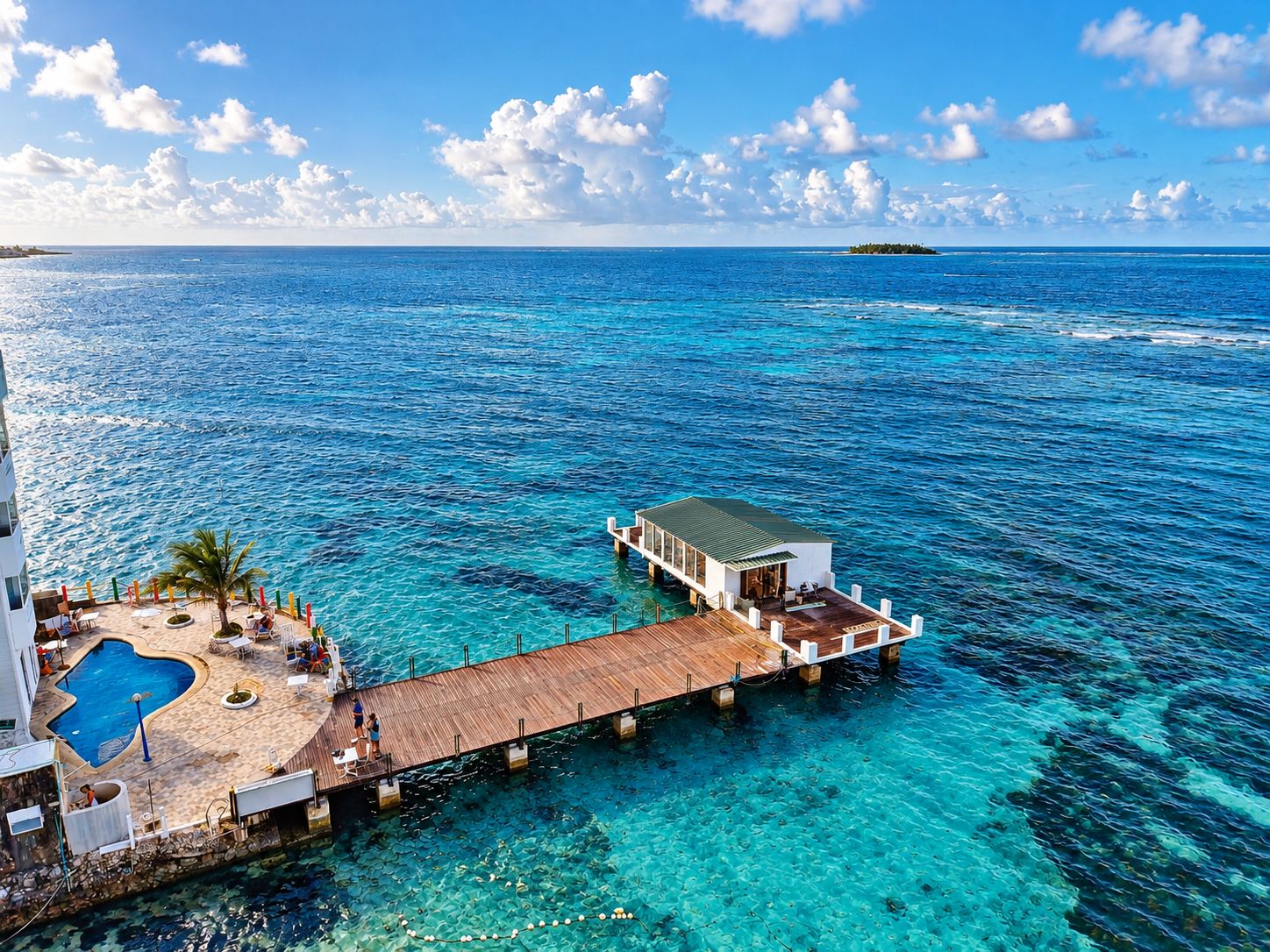 A resort with a wooden pier extending into clear blue waters, a pool area, and a small building with a green roof.