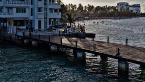 Un hotel frente al agua con un muelle que se adentra en el agua, un edificio con balcones y palmeras al fondo.