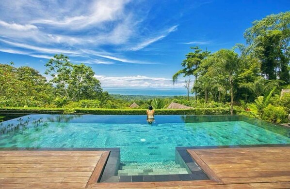 Vista despejada de una piscina infinita con una terraza de madera al frente, rodeada de exuberante vegetación y un cielo azul claro.