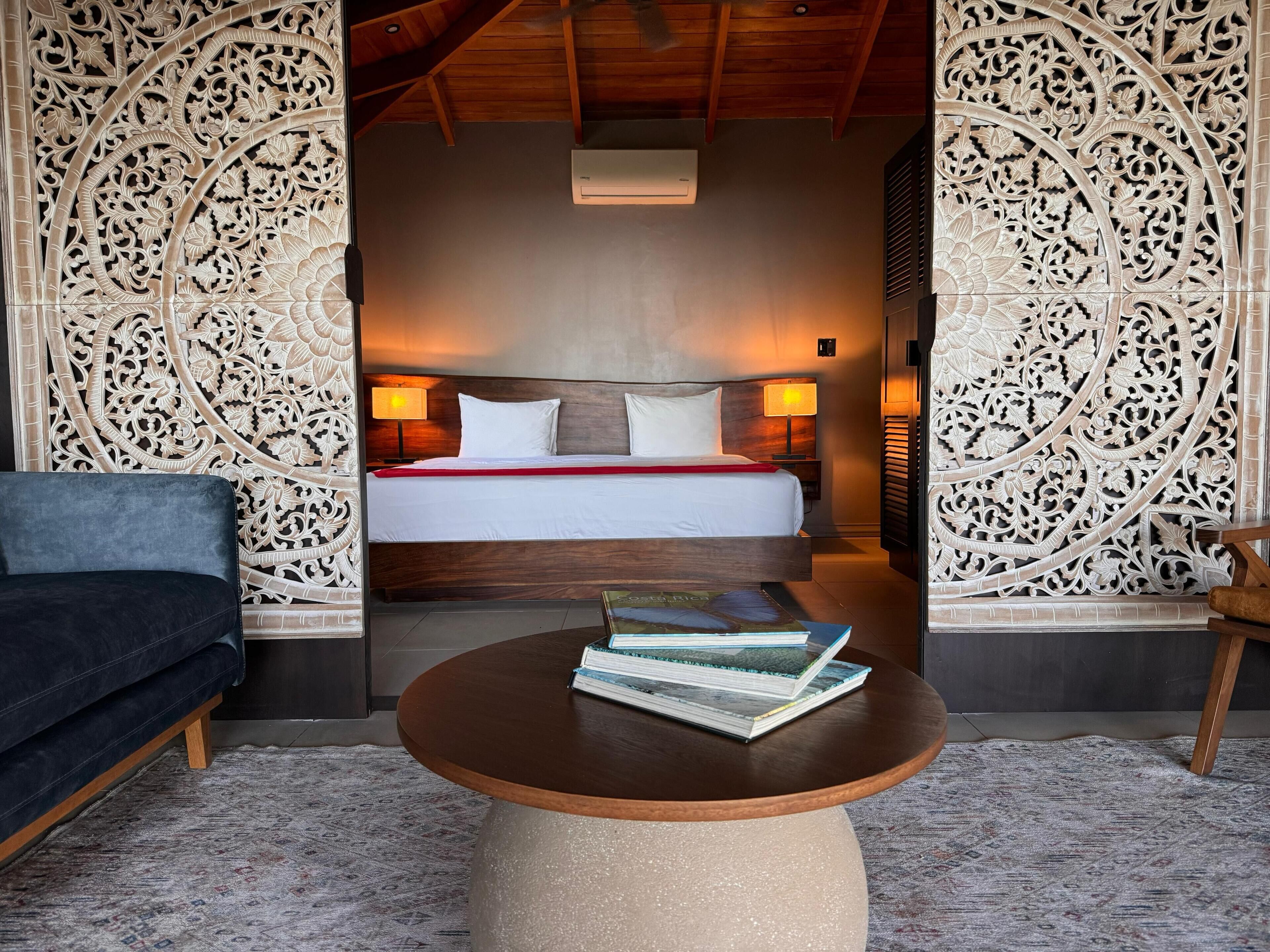 A hotel room with a bed, two bedside lamps, a round coffee table with books, a dark blue sofa, and intricate wall carvings.