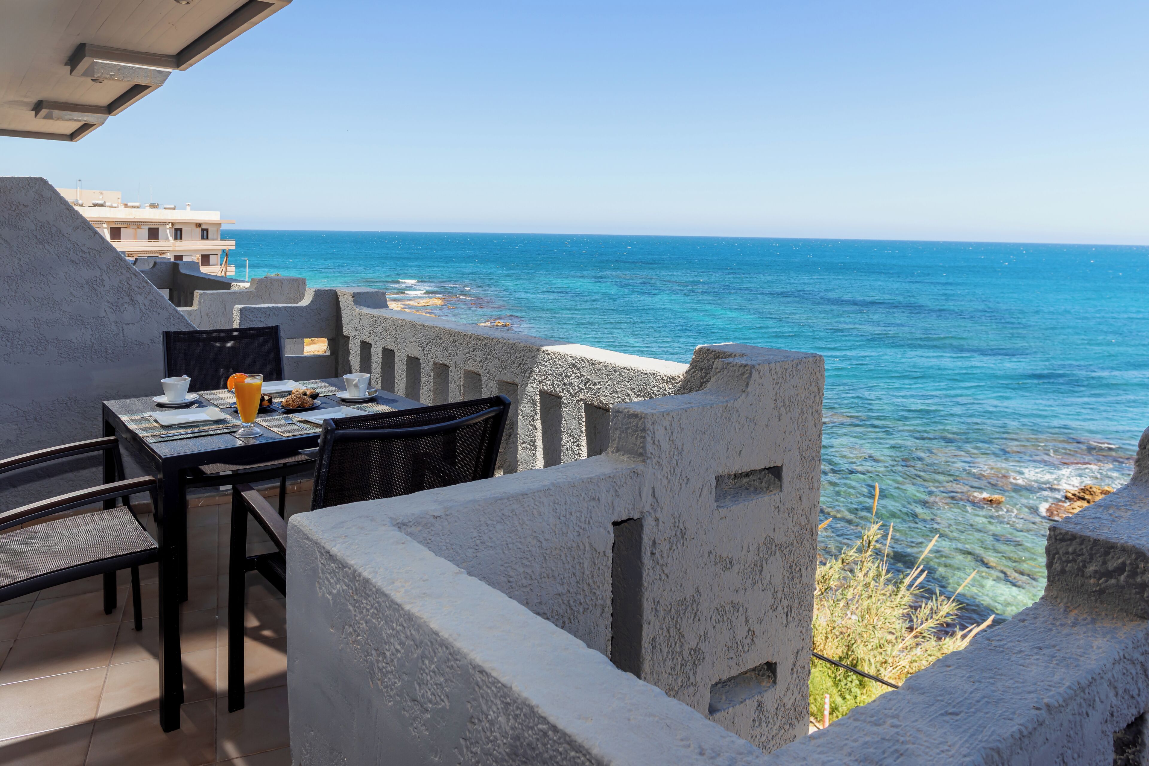 A balcony with a table set for two, overlooking a clear blue sea and rocky coastline.