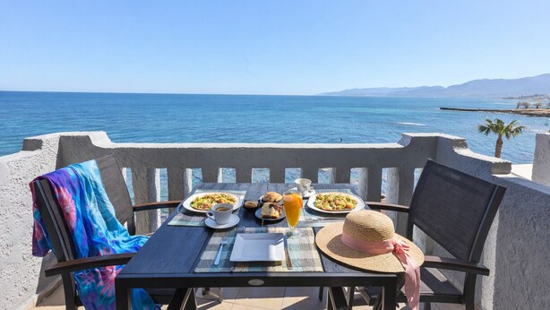 Une table dressée avec des assiettes remplies de nourriture, une boisson et un chapeau de paille, donnant sur une étendue d’eau avec des montagnes à l’horizon.