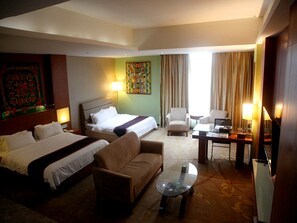 A hotel room with two beds, a sofa, a glass coffee table, and a desk with a lamp.
