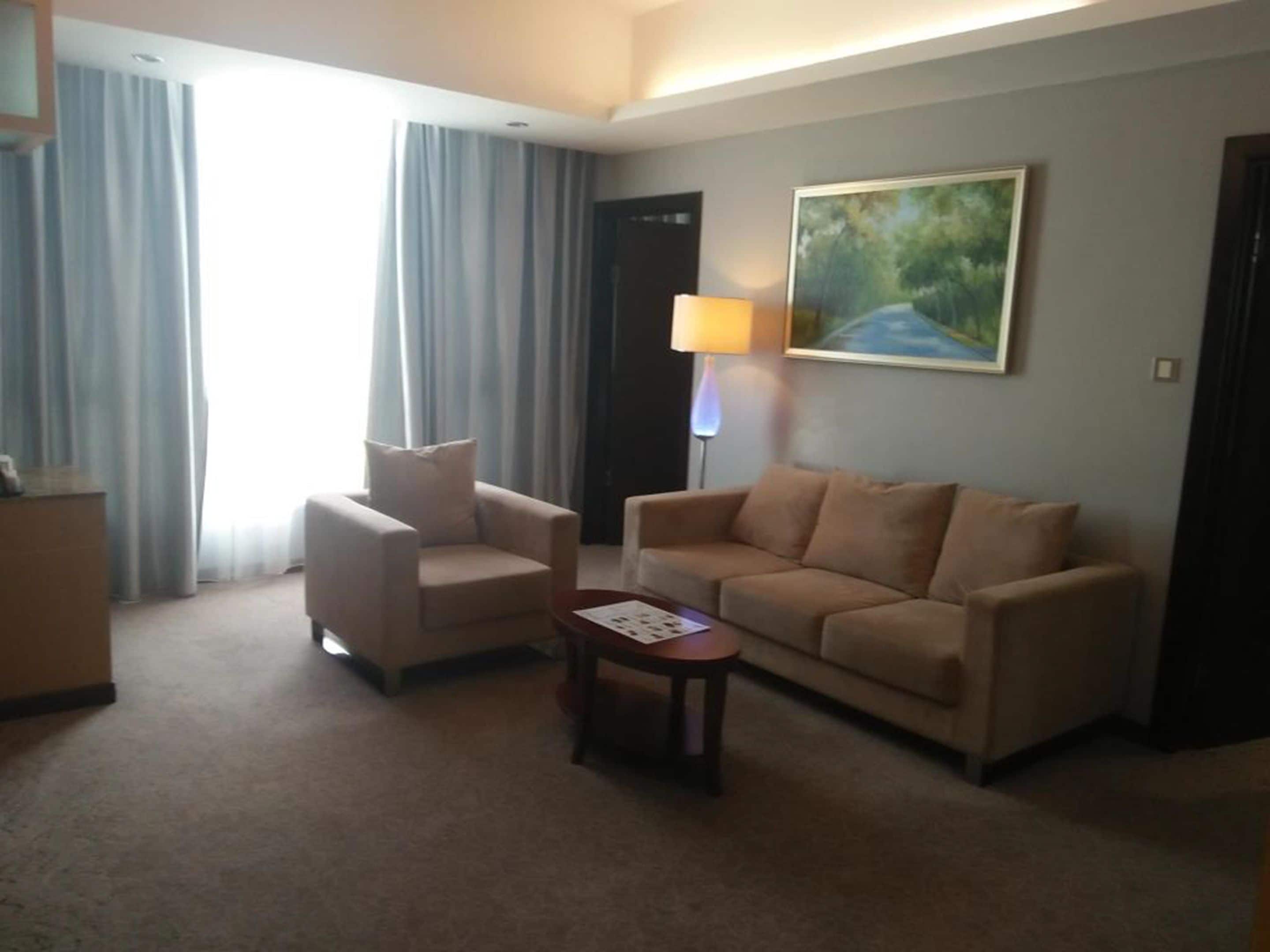 Ett hotellrum med en beige fåtölj, en beige soffa, ett litet bord och en tavla på väggen.