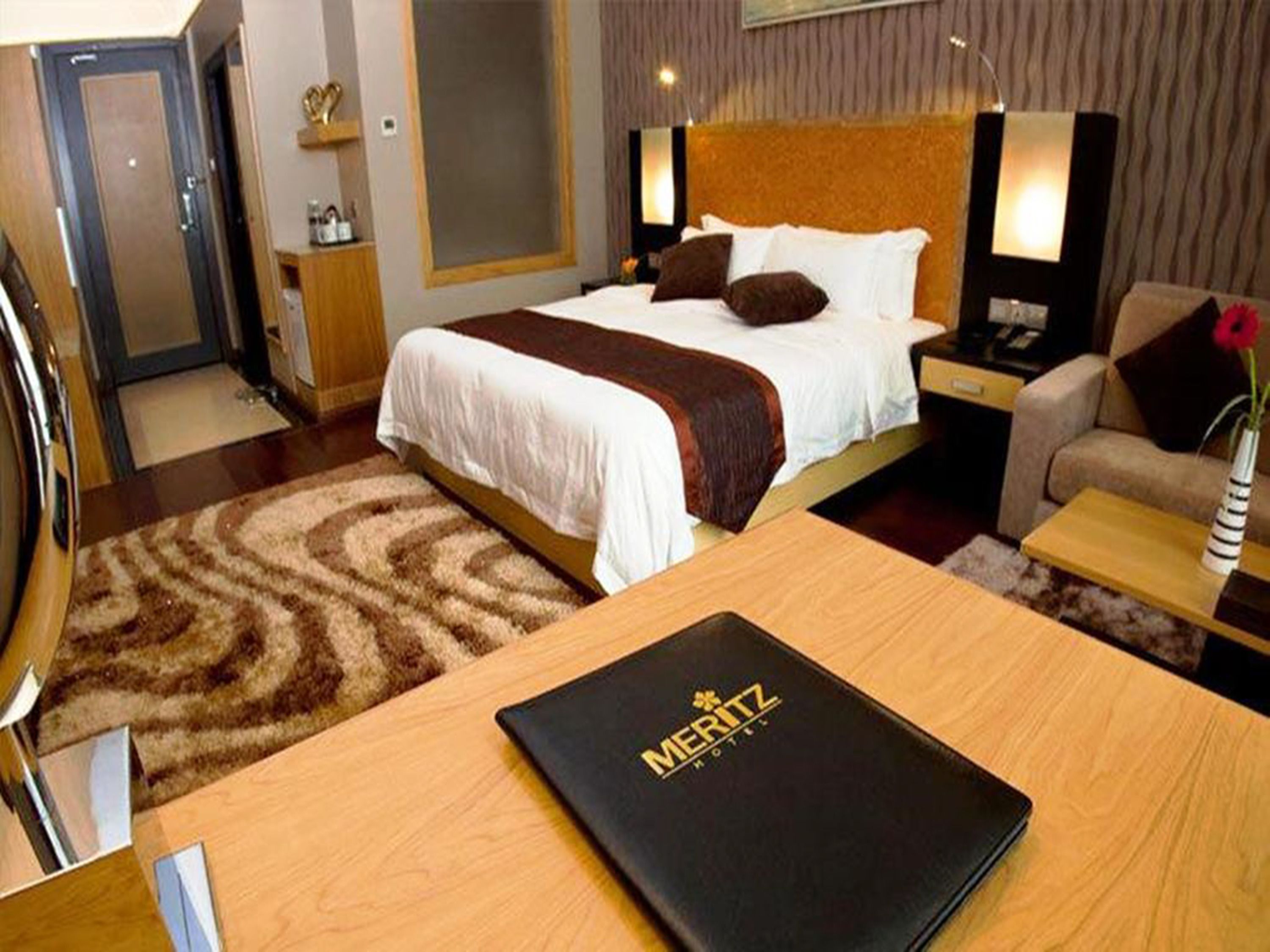 A hotel room with a bed, bedside tables, a desk, and a chair. There is a black folder with 'MERITZ HOTEL' on it on the desk.