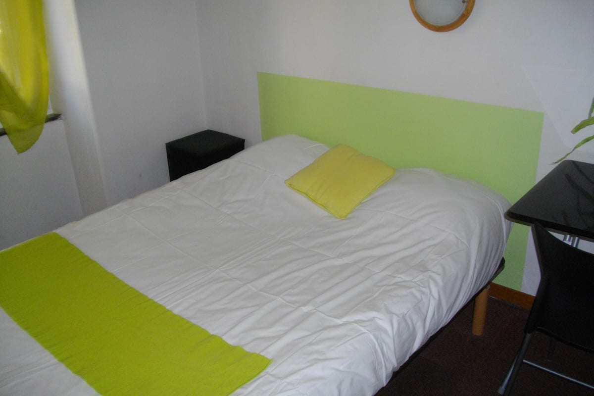 A single bed with a green and white bedspread, a yellow pillow, and a small bedside table.