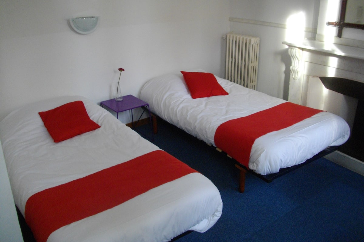 Two single beds with red pillows and a small purple table with a vase between them.