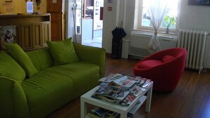 A room with a green sofa, a red chair, and a table with magazines. There is a window with curtains and a radiator. - Hotel Central (Carcassonne)