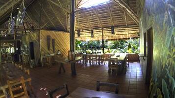 Un comedor interior con mesas y sillas de madera, techo de paja y vistas a exuberante follaje.