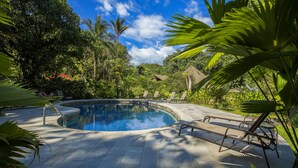 Uma piscina circular rodeada por espreguiçadeiras e vegetação tropical.